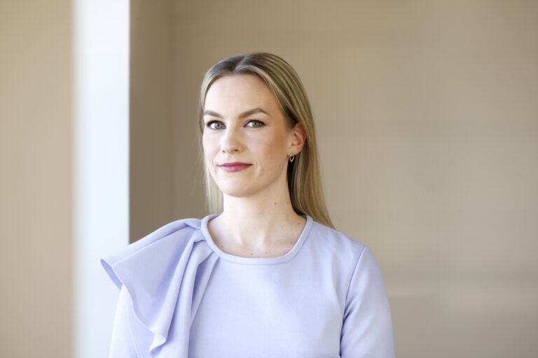 Vaalean lilaan mekkoon pukeutunut vaaleahiuksinen nainen kastoo kameraan ja hymyilee hiukan.